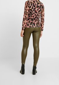 Langarm, figurbetontes Oberteil mit einem pinken und schwarzen Leopardenmuster, kombiniert mit olivgrünen, glänzenden Leggings und schwarzen Ankle Boots.