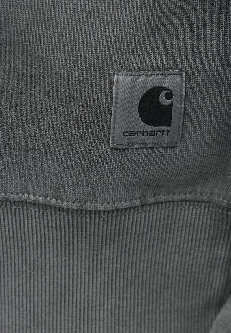 Sudadera gris con dobladillo acanalado, que presenta una etiqueta de tela cuadrada con un logo negro. Hecha de un material suave y texturizado, diseñada para la comodidad.