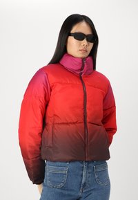 Giacca puffer color block rossa e rosa con colletto alto, chiusura a zip e due tasche frontali. Il tessuto appare liscio e gonfio.