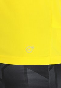 Gul fukttransporterande tröja med en strukturerad yta och en liten "DRY"-logotyp i ljusgrått vid fållen. Svarta mönstrade leggings delvis synliga.