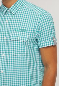 Camisa de manga corta con botones en patrón de cuadros vichy en verde azulado y blanco, con un bolsillo en el pecho y un logotipo bordado en la parte delantera.