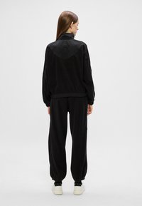 Femeie cu păr lung și brunet, îmbrăcată cu un jachetă și pantaloni negri de sport, stând cu spatele la cameră pe un fundal alb simplu.