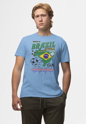 Jonge man met licht haar die een lichtblauw T-shirt draagt met het ontwerp van Brazilië, wereldkampioen voetbal 1994, met vlaggen en voetballen.