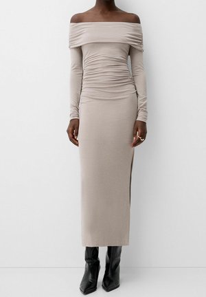 Robe longue à manches longues décolletée, en tissu beige clair avec détails froncés et fente latérale, associée à des bottes noires à bout pointu.