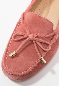 Mocassin en suédine rose avec un nœud noué et des embouts métalliques dorés sur fond blanc.