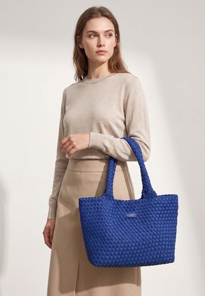 Donna che indossa top e gonna beige e tiene sulla braccio una grande borsa tote blu intrecciata con manici, su sfondo neutro.