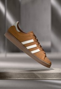 Rjavi1 semiš športni copat z belimi črtami in vezalkami, prikazan, kako lebdi nad kamnito platformo pred mehko osvetljenim sivo ozadjem.