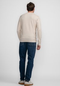 Beige sweater met lange mouwen en subtiele horizontale strepen, ronde hals, en geribbelde manchetten, gecombineerd met donkerblauwe jeans en witte sneakers.