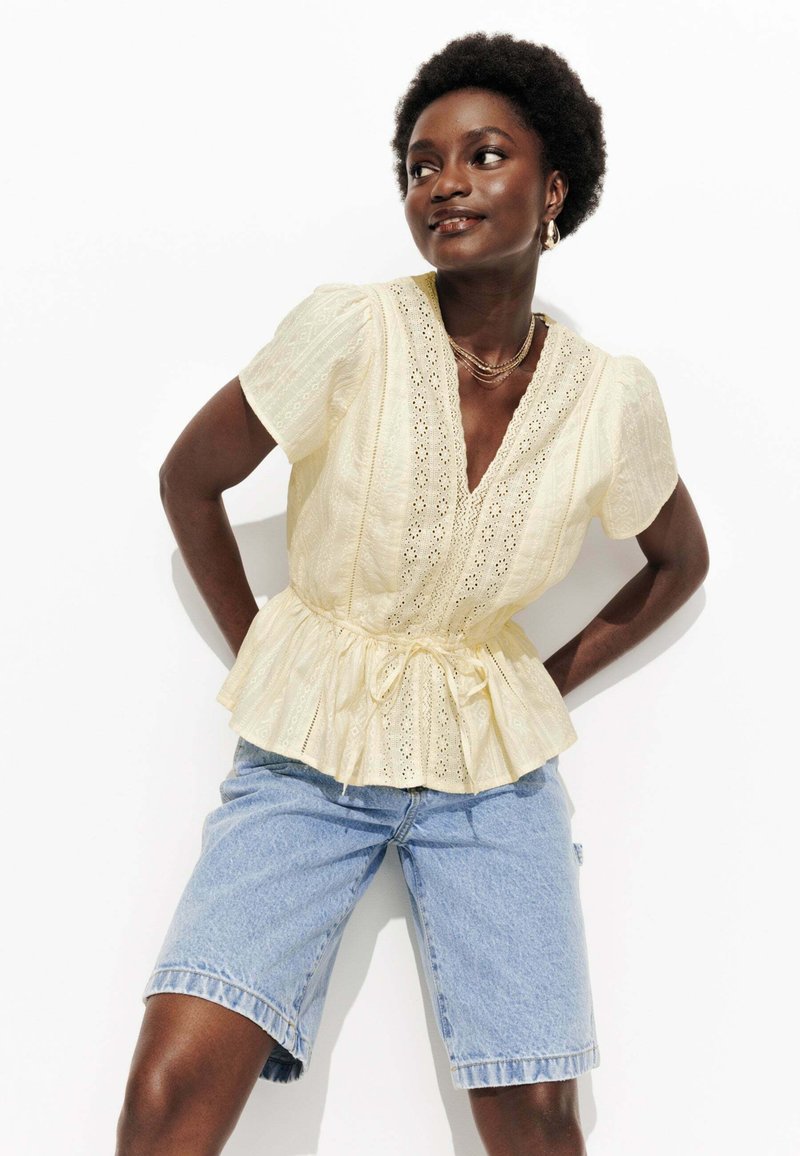 Blouse peplum jaune clair à manches courtes, avec des détails en dentelle et une taille à nouer. Associée à un short en denim bleu.