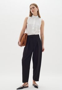 Vit ärmlös blus med hög krage, svarta skräddarsydda byxor och svarta spetsiga lägenheter. Håller i en brun läderväska. Minimalistisk design.