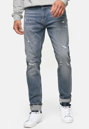 Homme portant un jean bleu clair délavé avec les revers roulés, assorti à des baskets blanches et un sweat-shirt gris, debout devant un fond blanc.