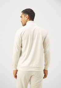 Svetlo béžová športová bunda s vysokým golierom, dlhými rukávmi a hladkým povrchom, spárovaná s rovnakými nohavicami pre súladný vzhľad.