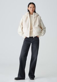 Cremefarbene Jacke mit strukturiertem Obermaterial, großen Taschen und Kapuze, getragen über einem leichten Pullover und kombiniert mit schwarzen, weiten Jeans.