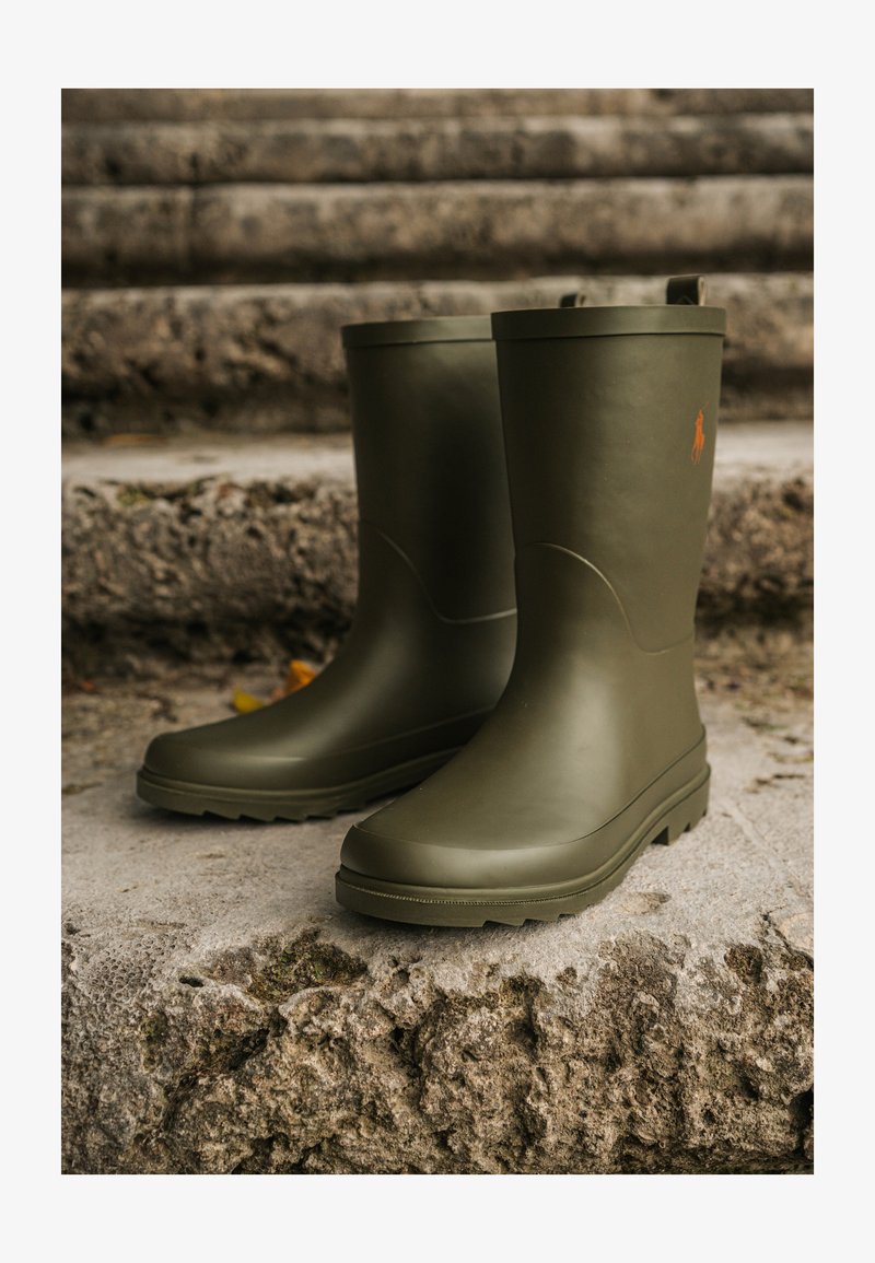 Stivali da pioggia in gomma verde con texture liscia e punta rotonda, caratterizzati da un logo arancione sul lato e una suola tassellata per una buona aderenza.