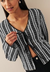 Cardigan a righe bianche e nere, con pizzo, scollo a V e chiusura con cravatta, dotato di maniche lunghe e una texture ad uncinetto intricata.