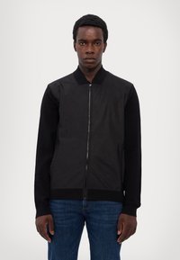 Black bomber jacket with a zip closure, contrasting knit sleeves, and a ribbed collar. Worn with blue denim jeans, featuring a smooth texture.