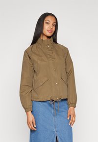 Giacca corta marrone con colletto alto, bottoni a pressione, tasche frontali e polsini elastici; indossata sopra una gonna di jeans blu.