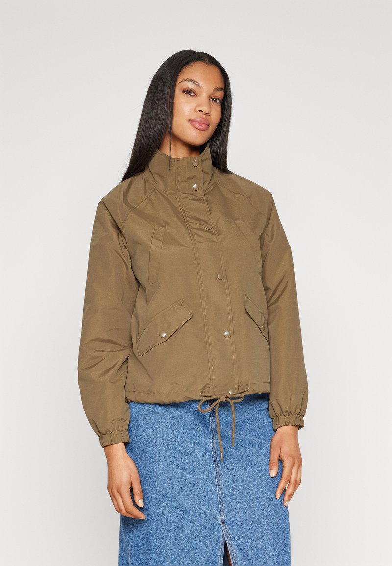 Giacca corta marrone con colletto alto, bottoni a pressione, tasche frontali e polsini elastici; indossata sopra una gonna di jeans blu.