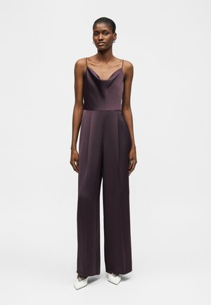 Γυναίκα που φοράει αμάνικο σκούρο μωβ σατέν ολόσωμο παντελόνι με φαρδιές φούστες και λαιμόκοψη καμπάνια, συνδυασμένο με λευκά μυτερά παπούτσια.