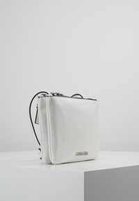 Biała skórzana torebka crossbody o prostokątnym kształcie, z zamkiem błyskawicznym i srebrnym akcentem logo. Posiada fakturowane wykończenie i regulowany pasek.