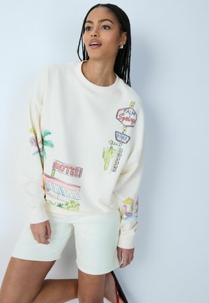 Frau mit geflochtenem Haar, die ein weißes Sweatshirt und Shorts mit bunten Vintage-Motel- und Diner-Schild-Illustrationen trägt.