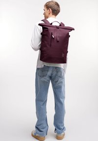 Maroon väskan med rulltopp, tillverkad av slitstarkt material; har justerbara remmar, en framficka med blixtlås och en logotyp i botten.