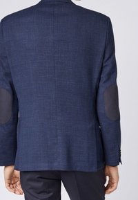 Marinblazer i texturerad tyg, med kontrasterande svarta elbow patches och en enda slits bak. Släta ytor och strukturerad design.