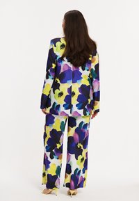 Femme aux longs cheveux bruns portant un tailleur floral coloré et des talons ouverts jaune, debout de dos sur fond blanc.