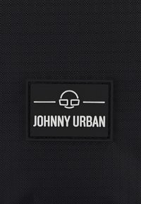 Černá texturovaná tkanina s obdélníkovou černou nášivkou s bílým nápisem "JOHNNY URBAN" a grafikou brýlí.
