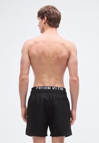 Μαύρα μαγιό βερμούδα από λείο ύφασμα, με λευκό λογότυπο "CALVIN KLEIN" στη ελαστική μέση και λεπτομέρεια πίσω τσέπης.