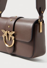 Bolso de hombro de cuero marrón con cierre en forma de pájaro dorado y etiqueta dorada grabada con el nombre de la marca "Pinko" en la correa.