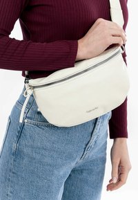 Sac banane en cuir blanc avec une finition texturée, fermeture éclair, et une sangle ajustable, porté sur un haut bordeaux et un jean bleu.