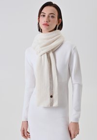 Sciarpa bianca sporca realizzata in un morbido tessuto testurizzato con delicate righe, caratterizzata da un piccolo bottone marrone e che si drappeggia elegantemente su un pullover bianco.