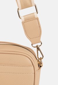 Esquina de bolso de cuero beige texturizado con cremallera dorada y cierre que sujeta una correa de tela ajustable a rayas crema y beige.