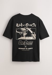 Fekete póló, amelyen egy monokróm illusztráció látható Monkey D. Luffyról, amint egy szalmakalapot hajt meg, rajta „One Piece” felirattal, angol és japán szöveggel.