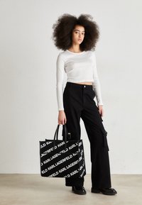 Svarta cargo-byxor och en kortärmad vit tröja med lång ärm, tillsammans med en stor svart tote-väska med vit logotyptext.