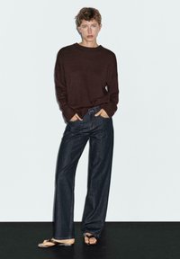 Brauner Pullover, lockere Passform, gerippte Bündchen; dunkelblaue weiter geschnittene Jeans mit klassischem Fünf-Taschen-Design; flache Sandalen mit minimalen Riemen.