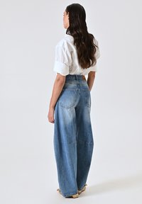 Jean denim bleu taille haute à jambes larges, avec des zones délavées et des poches standard. Associé à une chemise blanche ample.