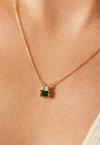 Gouden ketting met een smaragdvormige groene steen en een kleinere heldere steen, die een verfijnd ontwerp en fijne textuur weergeeft.