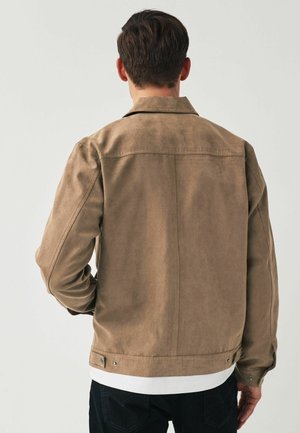 Muž v světle hnědém semišovém saku, bílém tričku a tmavých kalhotách, zobrazen zezadu na jednobarevném pozadí.