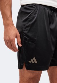 Άνδρας που φοράει μαύρα αθλητικά σορτς Adidas με πλαϊνή τσέπη και μικρό πλαϊνό σκίσιμο, στέκεται με το αριστερό χέρι χαλαρό δίπλα στο σώμα.