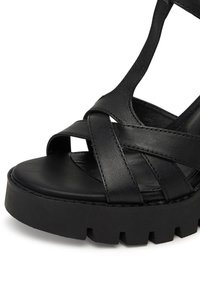 Sandal din piele neagră cu curele împletite, vârful deschis și o talpă platformă groasă, texturată, cu margini ondulate pentru aderență.