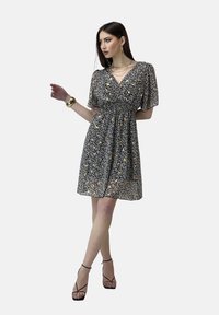 Vestido floral de manga corta con escote en V, base negra, patrones amarillos y blancos, cintura fruncida, hasta la rodilla, combinado con tacones con tiras.