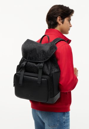 Jeune homme portant une veste rouge et un jean bleu, portant un grand sac à dos noir à motif avec rabat avant à boucle et poche zippée.