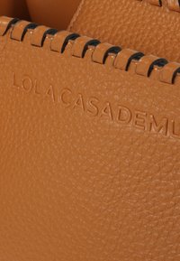 Oranžová kožená taška s texturou, která má prošitý černý okraj a embosované logo "LOLA CASADEMU." Povrch má vzor z kuliček.