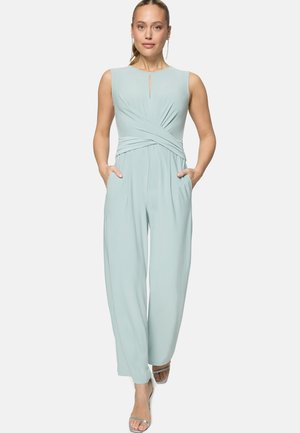 Woman walking forward wearing a sleeveless light blue jumpsuit with front pockets and silver open-toe heels on a white background.