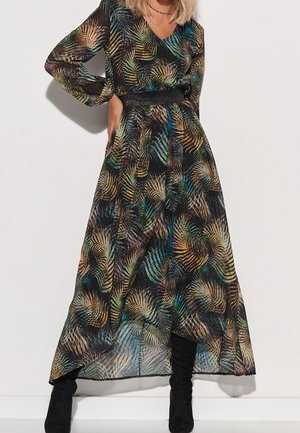 Femme portant une robe noire à manches longues, longue jusqu'aux chevilles, avec des motifs de feuilles multicolores et une ceinture noire scintillante à la taille, associée à des bottes noires.