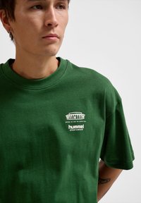 Green cotton t-shirt featuring a crew neckline and short sleeves. Front displays white printed logo and text. Smooth texture.