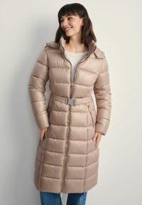 Beżowy, ocieplony płaszcz o długim kroju z paskiem w talii, zamek błyskawiczny i kaptur. Tkanina ma błyszczące wykończenie, poziome szwy i boczne kieszenie.