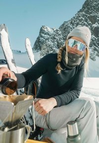 Eine Person in Winterkleidung gießt Kaffeemehl in eine silberne Kaffeemaschine. Im Hintergrund sind eine verschneite Landschaft und Skiausrüstung zu sehen.
